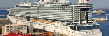 Sun Princess cruise ship docked at the port of Piraeus, with tugboats and ferries nearby.