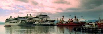 Cruise ships docked at the pier in Ushuaia harbour under a pink sky, with mountains in the distance.